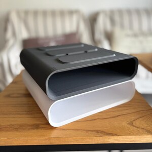 May include: A stack of two rectangular storage organizers with rounded corners. The top organizer is dark gray with three recessed rectangular indentations on the top surface, while the bottom organizer is a solid, smooth, translucent white.