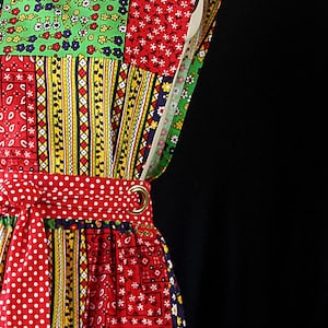 May include: A patchwork apron with a variety of colorful patterns, including polka dots, floral prints, and geometric designs. The apron features a red polka dot waist tie and pockets, and is displayed on a mannequin.
