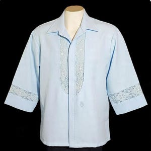 May include: Light blue short-sleeve shirt with white lace detailing on the front and sleeves. The shirt has a classic collar and a small embroidered design near the bottom. The shirt is displayed on a mannequin.