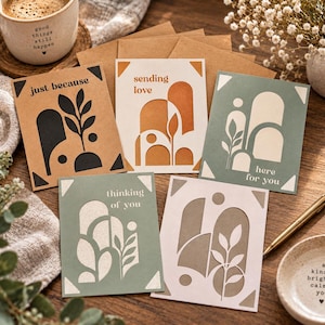 May include: Five rectangular greeting cards featuring abstract geometric shapes and botanical illustrations. The cards display phrases including just because, sending love, here for you, and thinking of you, printed in various colors on paper stock.