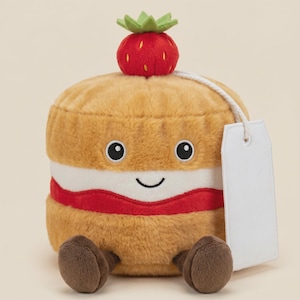 May include: A plush toy shaped like a round cake with a golden-brown exterior, a white cream layer, and a red jam filling. It features a smiling face, a red strawberry topper with green leaves, brown feet, and a blank white rectangular tag.