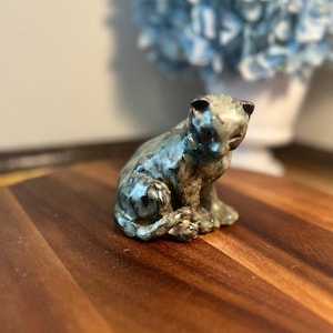 May include: A small, hand-sculpted ceramic figurine of a sitting cat. The piece features a mottled glaze with blended tones of deep blue, slate gray, and earthy brown, creating a textured, glossy finish across its rounded form and ears.