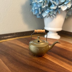 May include: A small brass teapot featuring a rounded body with etched semi-circular patterns, a curved spout, and a metal handle topped with a cylindrical wooden grip. The lid has a small central opening and a decorative cross-shaped vent.