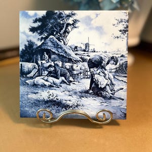 May include: Square ceramic tile with a blue and white pastoral scene depicting people shearing sheep near a thatched-roof building and a windmill. The tile is displayed on a decorative metal stand.