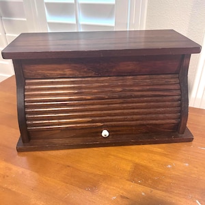 May include: A dark brown wooden breadbox with a roll-top door and a white knob. The breadbox has a rectangular shape with a flat top and a curved design on the sides. The wood grain is visible.