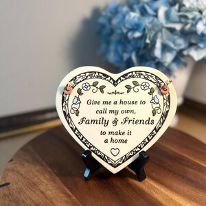 May include: Heart-shaped ceramic plaque with the text "Give me a house to call my own, Family & Friends to make it a home." The plaque has a black floral border and is displayed on a black stand. Two small pink accents are visible.