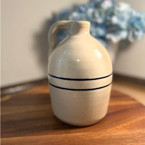 May include: A cream-colored ceramic jug with a single handle and a narrow neck. The body of the jug features two distinct, parallel dark blue horizontal stripes encircling the midsection. The surface has a smooth, glossy finish throughout.