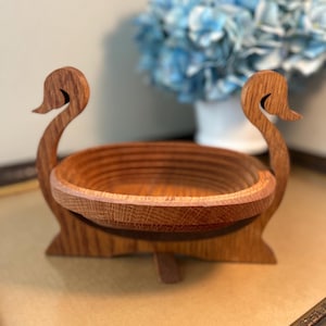 May include: A wooden swan-shaped fruit bowl. The bowl is made of light brown wood and features a round, slatted design. Two swan heads form the handles, and the bowl sits on a base with a small support. The background includes a vase of blue flowers.