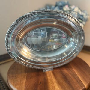 May include: An oval silver-toned metal serving tray featuring a polished, reflective center surface and a decorative rope-style textured border along the outer rim. The tray is positioned upright, showcasing its smooth, metallic finish and oval shape.