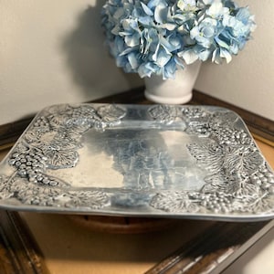 May include: A square silver-toned metal serving tray featuring a raised, embossed border design of grape clusters and vine leaves. The center of the tray is smooth and reflective, while the edges display detailed botanical textures.