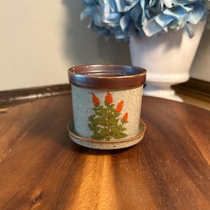 May include: A small, speckled ceramic planter with a matching saucer. The planter features a brown rim and a green and orange floral design. The planter is sitting on a wooden surface, with a white vase of blue flowers in the background.