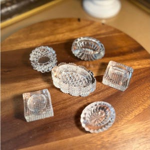 May include: A collection of six clear glass items including a circular ring, two square-based pieces with circular indentations, two oval-shaped dishes with textured edges, and one central oval dish featuring a detailed cross-hatched pattern.