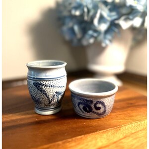 May include: A tall, cylindrical ceramic cup and a shorter, wide ceramic bowl, both featuring a light gray base with dark blue hand-painted patterns. The cup has a flared rim and a textured dotted design, while the bowl displays a swirling blue motif.
