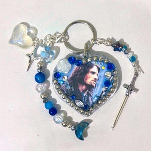 Peut inclure: Un porte-clés en forme de cœur avec un portrait d'homme aux cheveux longs, orné de perles bleues et argentées, d'un petit charme d'épée et d'un croissant de lune. Le porte-clés a un anneau argenté et un charm en forme de cœur transparent.