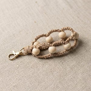 May include: A macrame keychain with a natural, woven design. The keychain features a series of light-colored wooden beads and a gold-colored metal clasp. The macrame is a light brown color, and the beads are round.