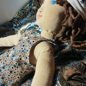 Pode incluir: Boneca feita à mão com corpo de tecido bege, cabelo de fio castanho e olhos bordados azuis. A boneca usa um colete estampado com círculos azuis, castanhos e bege. Um lenço branco com um desenho azul cobre a cabeça. A boneca repousa sobre um tecido estampado.