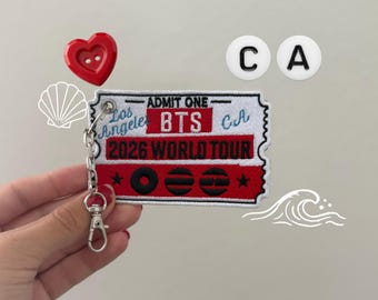 Puede incluir: Un parche bordado rectangular con forma de boleto y un llavero de metal adjunto. El parche presenta el texto ADMIT ONE, Los Angeles, BTS, CA y 2026 WORLD TOUR en hilo rojo, negro y azul sobre un fondo blanco con bordes de color rojo.