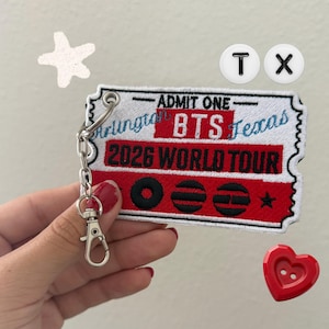 May include: A rectangular embroidered keychain shaped like a ticket with white, red, and black thread. It features the text ADMIT ONE, BTS, Arlington, Texas, and 2026 WORLD TOUR. The keychain includes a silver metal chain and a lobster clasp.