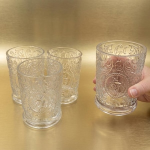 May include: Four clear, ornate drinking glasses with a detailed embossed pattern. One glass is held by a person. The glasses are on a gold surface.