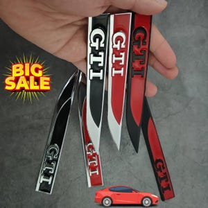 May include: A collection of GTI car emblems in black, red, and silver, held against a gray background. The emblems are long and narrow with the GTI logo in white. A small red car is at the bottom. A "BIG SALE" sign is in the upper left corner.