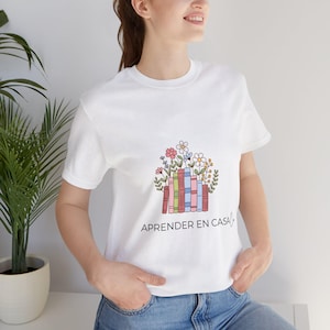 May include: White t-shirt with a colorful graphic of books and flowers. The design includes the text "APRENDER EN CASA" in black. The shirt is made of a soft material and has short sleeves.