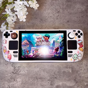 Könnte beinhalten: Eine weiße tragbare Spielkonsole mit einer individuellen Hülle, die bunte Cartoon-Illustrationen von Süßigkeiten wie Eis, Bonbons und Zuckerwatte zeigt. Auf dem Bildschirm ist der Spieltitel Discounty mit animierten Charakteren zu sehen.