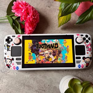 May include: A white handheld gaming console with a screen displaying the game "Eastward." The console is decorated with cartoon stickers. A pink flower and green plants are in the background.