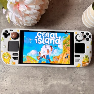 Puede incluir: Una consola de juegos portátil blanca con una carcasa personalizada que incluye varias pegatinas pequeñas, como una rana amarilla, un caracol y un pato. La pantalla muestra el título del juego Coral Island. Los joysticks tienen fundas de oso marrón y conejo blanco.