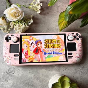 May include: A pink handheld games console displaying "Story of Seasons: Grand Bazaar". The console has a pink case with decorative stickers and buttons. The background includes white flowers and green plants. The console is on a grey surface.