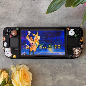 May include: A black handheld gaming console featuring a screen displaying a pixel art scene of a person riding a donkey. The console is decorated with various stickers including a ghost, a cat, a bear, and a rabbit, along with small stars and moons.