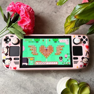 May include: A pink handheld gaming console with a game displayed on the screen. The console is decorated with stickers of a cartoon character and hearts. A pink flower and green plants are in the background.