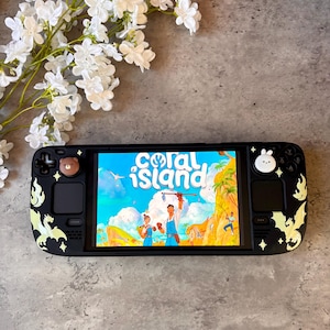 May include: A black handheld gaming console featuring a screen displaying the Coral Island game title. The device is decorated with light-colored dragon decals, small star accents, and two thumbstick covers shaped like a brown bear and a white rabbit.