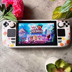 May include: A white handheld gaming console with a screen showing a colourful game scene. The console is decorated with peach and flower designs. The game title "Discounty" is visible on the screen. Pink flowers and green plants surround the console.