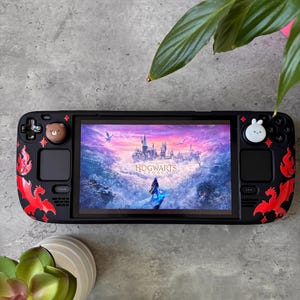 Könnte beinhalten: Eine schwarze Handheld-Spielkonsole mit einer individuellen Schutzfolie, die rote Drachenmotive und kleine Sternakzente zeigt. Auf dem Bildschirm ist der Startbildschirm von Hogwarts Legacy zu sehen. Die Joysticks haben einen Bären- und Hasenaufsatz.