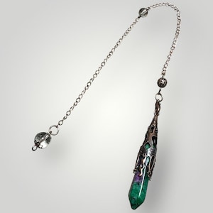 May include: A pendulum with a silver chain and a faceted crystal at the end. The pendulum features a green and purple crystal encased in a decorative metal cage. The chain has a small crystal bead and a decorative metal bead.