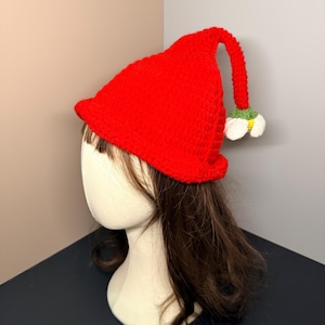 May include: A bright red, crocheted hat with a whimsical design. The hat features a long, curved stem ending in a white and green flower. The hat is displayed on a mannequin head with brown hair, showcasing its unique shape and style.