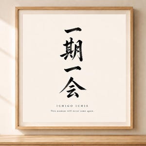 以下が含まれることがあります： ライトウッドフレームに入った正方形のウォールアートプリント。中央に縦書きの黒い日本語の書道文字「一期一会」が描かれています。文字の下には「ICHIGO ICHIE」および「This moment will never come again.」というテキストが記載されています。