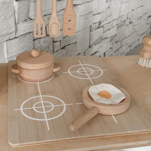 May include: Wooden toy kitchen set with a stove, pot, pan with a fried egg, and utensils. The stove has white markings for burners. A carton of wooden eggs sits above the stove. The set is made of light-colored wood.