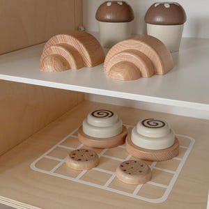以下が含まれることがあります： 木製の玩具セット。虹のアーチ2つ、キノコ型のカップ2つ、シナモンロール2つ、クッキー2つ。おもちゃは天然木で作られ、ニュートラルカラーで塗装されており、想像力豊かな遊びに最適です。