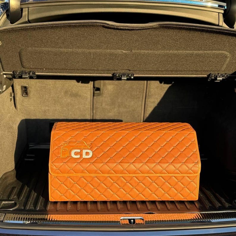 Op de afbeelding: Een oranje, gewatteerde auto-kofferbakorganizer met de letters "CD" op de voorkant. De organizer is rechthoekig en heeft een ruitpatroon. De kofferbak is open en onthult de organizer binnenin. Het interieur van de auto is zwart.