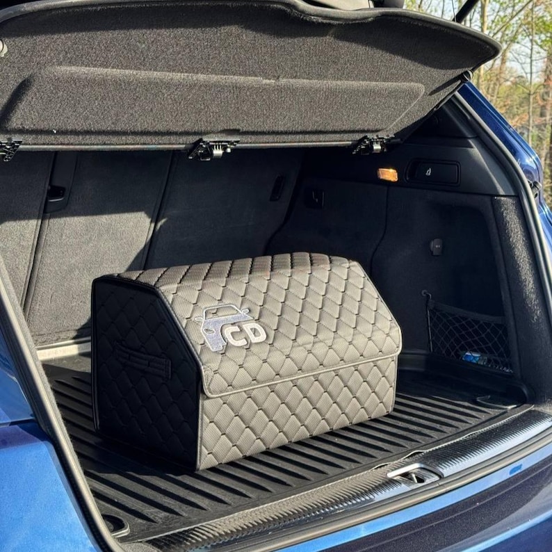 K&ouml;nnte beinhalten: Ein schwarzer, gesteppter Kofferraum-Organizer mit Rautenmuster und einem Autologo mit den Buchstaben "CD" auf der Vorderseite. Der Organizer befindet sich im Kofferraum eines blauen Autos.