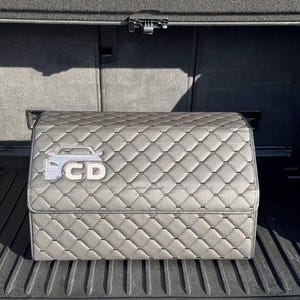 K&ouml;nnte beinhalten: Grauer, gesteppter Kofferraum-Organizer mit silberner Auto-Umrisslinie und den Buchstaben "CD". Der Organizer ist rechteckig mit einem perforierten Muster und einem Rautenstich-Design. Der Organizer ist f&uuml;r den Kofferraum eines Autos konzipiert.