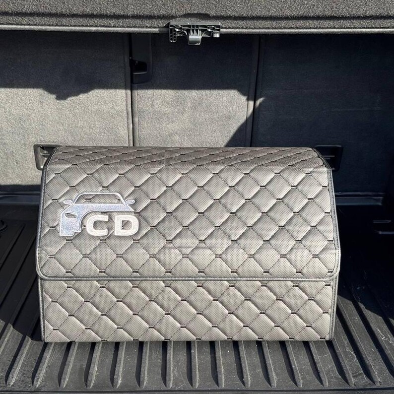 K&ouml;nnte beinhalten: Grauer, gesteppter Kofferraum-Organizer mit silberner Auto-Umrisslinie und den Buchstaben "CD". Der Organizer ist rechteckig mit einem perforierten Muster und einem Rautenstich-Design. Der Organizer ist f&uuml;r den Kofferraum eines Autos konzipiert.