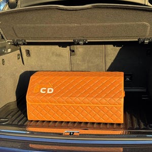 Op de afbeelding: Een oranje, gewatteerde opbergcontainer met de letters "CD" op de voorkant. De container is rechthoekig en lijkt gemaakt van een leerachtig materiaal. Hij staat in de kofferbak van een voertuig.
