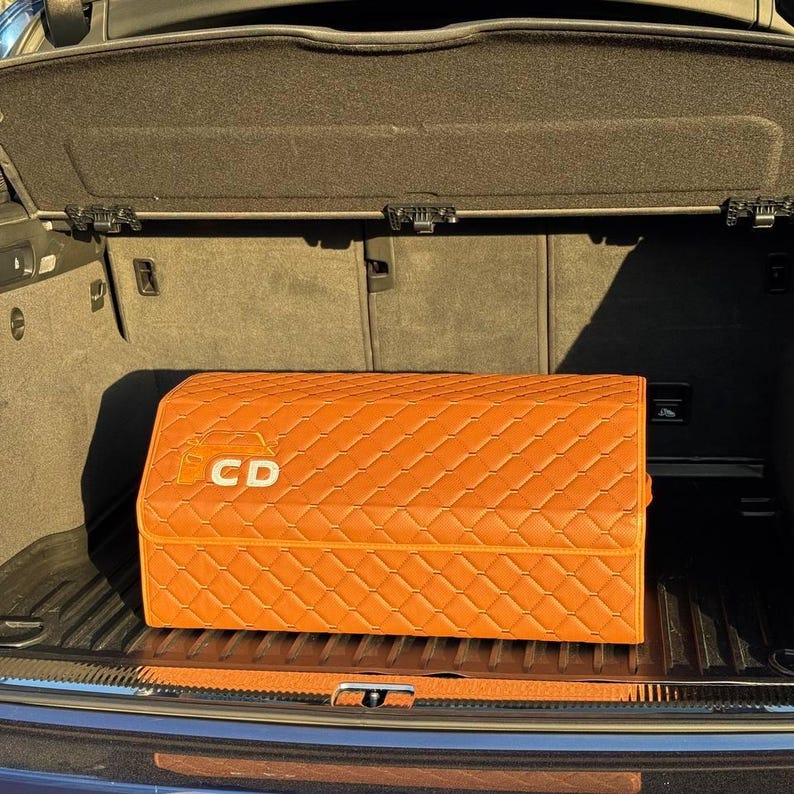 Op de afbeelding: Een oranje, gewatteerde opbergcontainer met de letters "CD" op de voorkant. De container is rechthoekig en lijkt gemaakt van een leerachtig materiaal. Hij staat in de kofferbak van een voertuig.