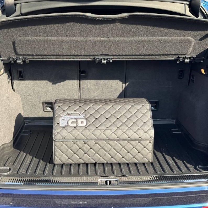 K&ouml;nnte beinhalten: Ein grauer, gesteppter Kofferraum-Organizer mit Rautenmuster und einem Auto-Logo mit den Buchstaben "CD" auf der Vorderseite. Der Organizer befindet sich in einem Kofferraum.