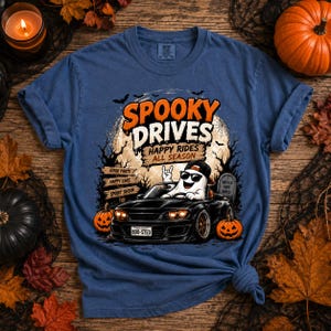 May include: A blue short-sleeve t-shirt featuring a graphic of a ghost wearing sunglasses and a cap driving a black sports car. The text reads SPOOKY DRIVES HAPPY RIDES ALL SEASON, with pumpkins, a full moon, and a wooden signpost in the background.