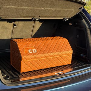 Op de afbeelding: Een oranje, gewatteerde auto-kofferbakorganizer met een ruitpatroon. De rechthoekige organizer heeft de letters "CD" op de voorkant. Hij staat in de kofferbak van een auto.