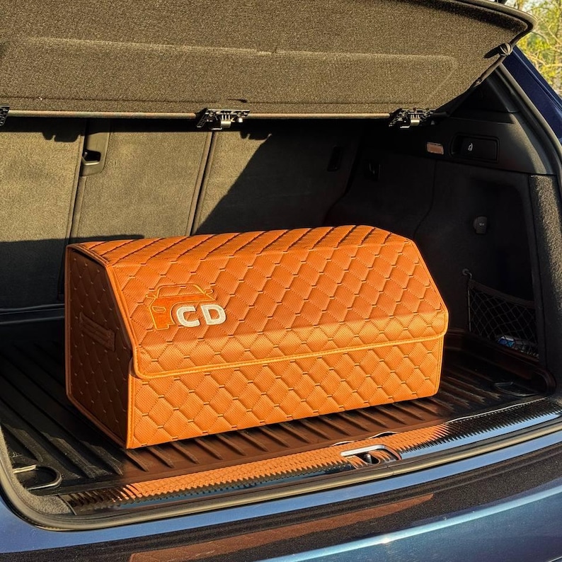 Op de afbeelding: Een oranje, gewatteerde auto-kofferbakorganizer met een ruitpatroon. De rechthoekige organizer heeft de letters "CD" op de voorkant. Hij staat in de kofferbak van een auto.