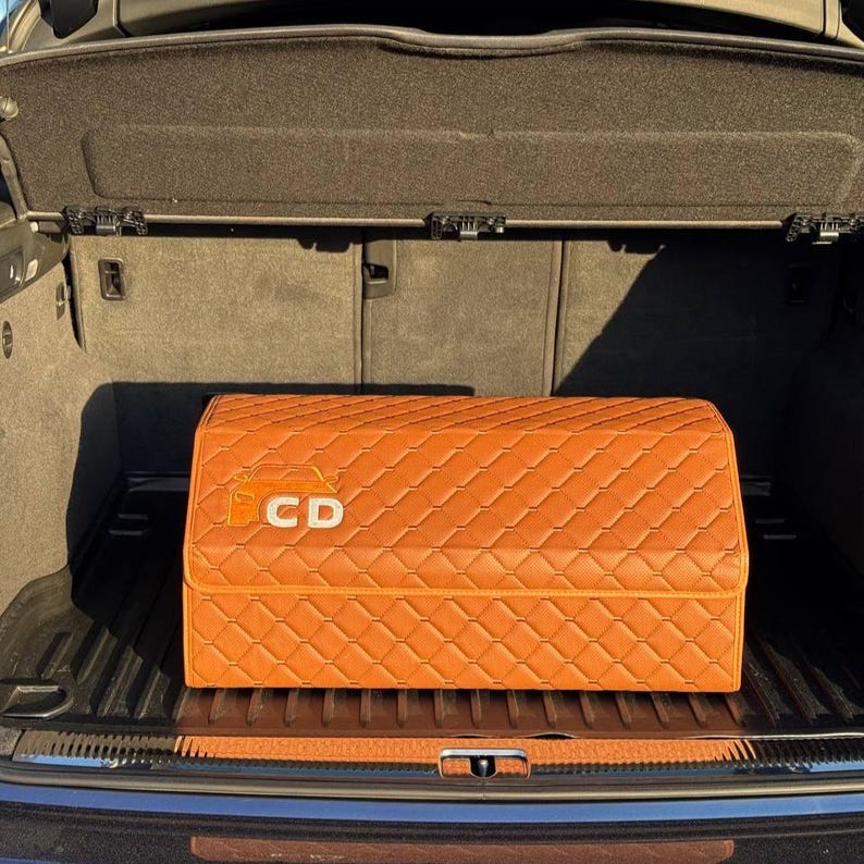Op de afbeelding: Een oranje gewatteerde opbergorganizer met de letters "CD" in het wit, geplaatst in de kofferbak van een auto. De organizer heeft een ruitpatroon en een rechthoekige vorm. De kofferbak is donkergrijs.